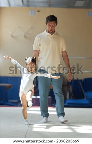 Full shot of cheerful Asian little girl wearing white bodysuit exercising in gymnastics class while Caucasian male coach following and supervising her Royalty-Free Stock Photo #2398203669