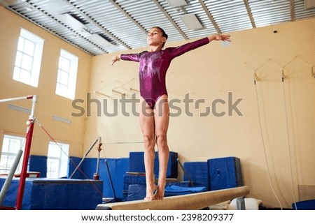 Full shot of confident young girl wearing pink leotard standing on toes with hands in air balancing on beam in gymnasium, copy space Royalty-Free Stock Photo #2398209377