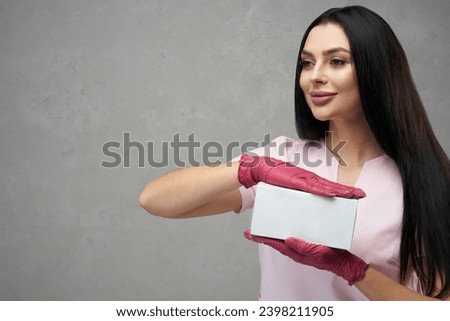 Happy female cosmetologist holding white box with both hands. Portrait of dark haired pretty woman wearing pink gloves and uniform, showing small box against grey wall. Beauty, cosmetology concept. Royalty-Free Stock Photo #2398211905