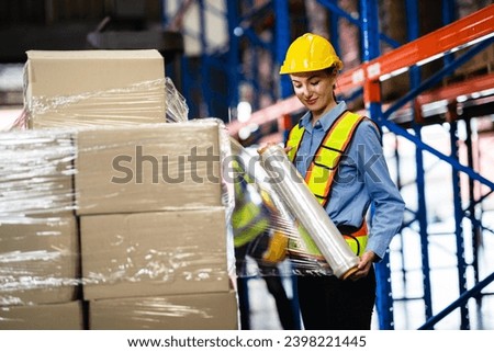 Female Caucasian warehouse worker wearing a hard hat and reflective jacket wraps boxes in stretch film parcels on pallets while controlling stock. Warehouse concept in retail distribution center