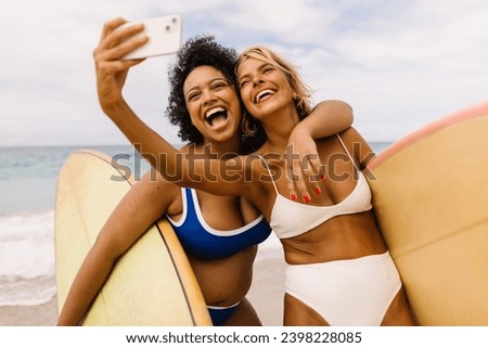 Best friends on a surfing vacation capture the moment with a joyful selfie on the beach, their smiles reflecting the exciting memories they create. Happy female surfers having fun together on shore.