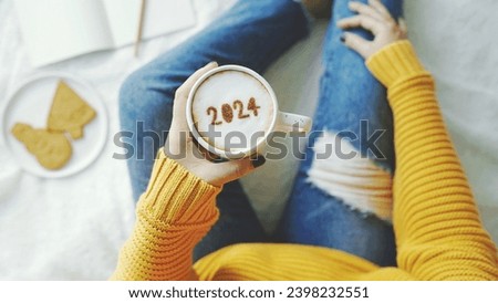 Holidays food art number 2024 on frothy surface of cappuccino in coffee cup holding by woman in yellow knitted sweater with jeans sitting on bed with white blanket background. New Year's goal setting.