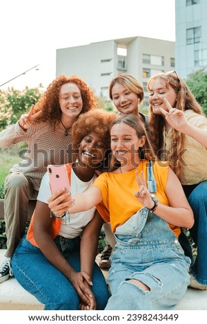 Vertical. Happy multiracial young women looking at camera with a smart phone enjoying together sitting outside. Funny ladies having fun shooting a selfie portrait their friends with cellphone. High