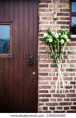 This inviting scene features a floral arrangement of white roses and complementary greenery, standing tall in a vase beside a rich wooden door. The rustic charm of the brick wall enhances the beauty