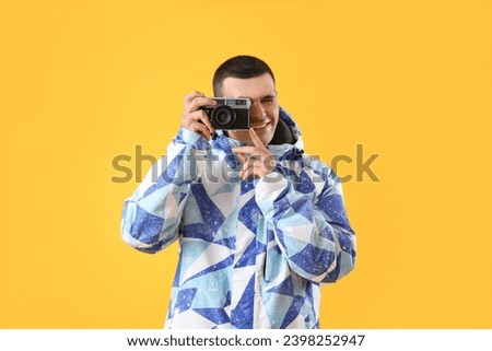Young man in warm jacket with photo camera on yellow background