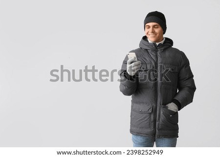 Young man in puffer jacket using mobile phone on light background
