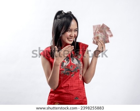 Portrait of a happy Asian woman wearing a traditional red Cheongsam Qipao dress, showcasing a stack of money, isolated against a white background.