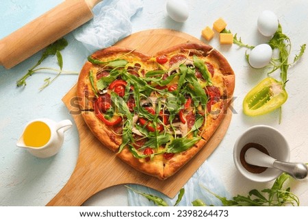Wooden board with tasty heart shaped pizza and ingredients on light blue background