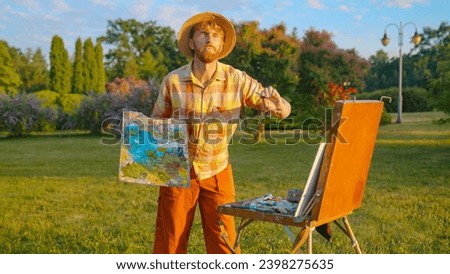 The artist paints a picture in the park. Green landscape, grass trees and flowers. The easel on which the painting stands.