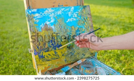 The artist paints a picture in the park. Green landscape, grass trees and flowers. The easel on which the painting stands.