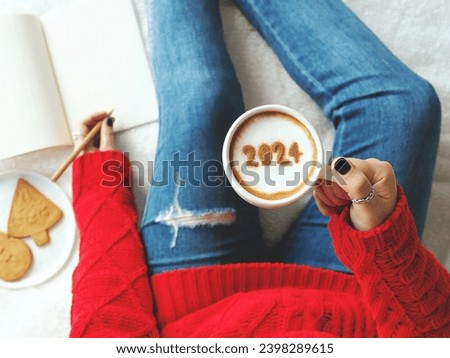 2024 New Year's goal setting, number 2024 on frothy surface of cappuccino in white coffee cup holding by woman in red knitted sweater with jeans sitting on bed while writing down her resolutions. Royalty-Free Stock Photo #2398289615
