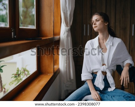 Woman sitting at home by a wooden window with a smile on a chair in homemade comfortable clothes and looking at the landscape, spring mood, women's day, rest on the weekend. Royalty-Free Stock Photo #2398308077