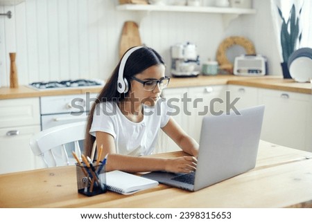 Pre-teen 12s girl in headphones and eyeglasses sit at table e-learns, listen online course, audio lesson, get new knowledge, skills using internet and modern tech. Development, education, eye-sight Royalty-Free Stock Photo #2398315653