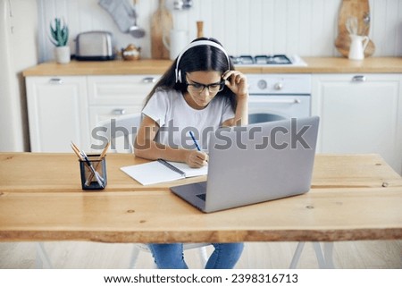 Pre-teen 12s girl in headphones and eyeglasses sit at table e-learns, listen online course, audio lesson, get new knowledge, skills using internet and modern tech. Development, education, eye-sight Royalty-Free Stock Photo #2398316713