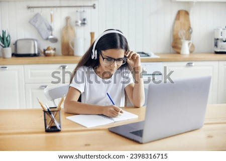 Pre-teen 12s girl in headphones and eyeglasses sit at table e-learns, listen online course, audio lesson, get new knowledge, skills using internet and modern tech. Development, education, eye-sight Royalty-Free Stock Photo #2398316715