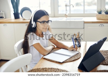 Pre-teen 12s girl in headphones and eyeglasses sit at table e-learns, listen online course, audio lesson, get new knowledge, skills using internet and modern tech. Development, education, eye-sight Royalty-Free Stock Photo #2398316719