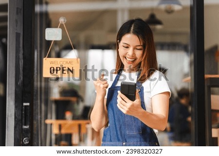 Happy excited attractive young Asian woman in denim apron, received online order in coffee shop with an 'Open' sign.