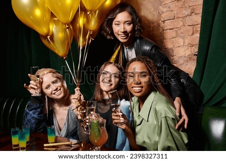 joyful multiracial women with champagne looking at camera near golden balloons in bar, birthday