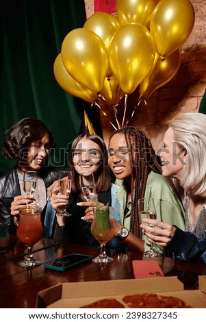 happy woman in party hat with champagne near multiethnic girlfriends and birthday balloons in bar