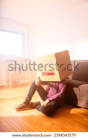 Full body of faceless male sitting on parquet floor leaning on sofa with cardboard box with square eyes and rectangular mouth on top of head on sunny day at home