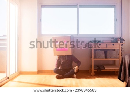 Full body of faceless male sitting on parquet floor leaning on a wall while hiding head in cardboard box with square eyes and rectangular mouth on sunny day