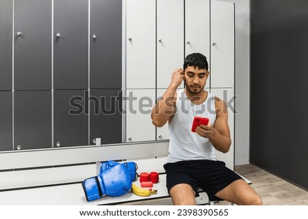 Horizontal photo man young adult mixed race sitting in the van of the boxing gym locker room with his cell phone in his hand and placing headphones in his ear. Copy space. Sports, technology.
