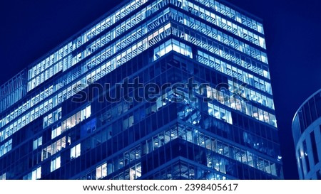Fragment of the glass facade of a modern corporate building at night. Big glowing windows in modern office buildings at night, in rows of windows light shines. Blue graphic filter. Royalty-Free Stock Photo #2398405617