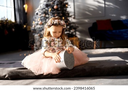 child girl sitting in front of a Christmas tree in a pink dress and opening a gift