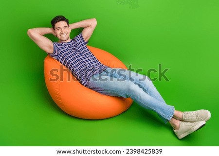 Full size photo of positive guy dressed striped t-shirt denim pants hold hands behind head lay on pouf isolated on green color background