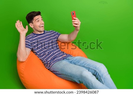 Full size photo of nice guy dressed t-shirt pants sit on pouf waving hand on smartphone make selfie isolated on green color background