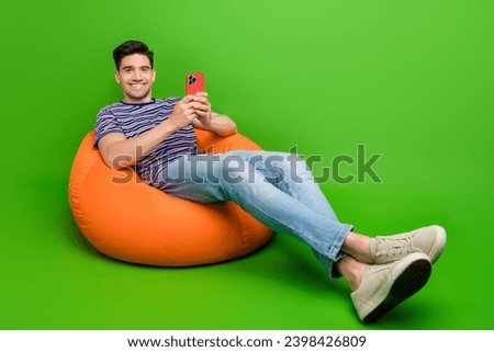 Full size photo of good mood guy dressed t-shirt denim pants sit on pouf read news on smartphone isolated on green color background