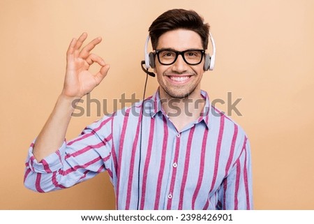 Photo of cheerful call center operator in wired headset young man showing okey sign wear striped shirt isolated on beige color background Royalty-Free Stock Photo #2398426901