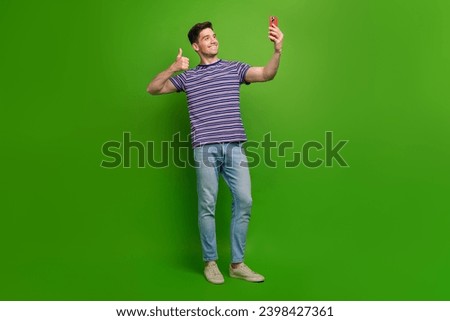 Full size photo of good mood guy dressed striped t-shirt showing thumb up make selfie on smartphone isolated on green color background