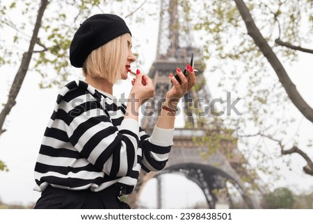 Stylish blonde in fashionable black beret paints her lips with red lipstick, against background of Eiffel Tower in Paris on spring day. Walking aroun dcity. Beauty and fashion. Tourism and travel. 