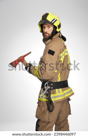 Serious caucasian firefighter in helmet holding big axe in studio. View from shoulder of bearded male rescuer with hatchet, looking at camera, on gray background. Concept of job, fireman equipment.