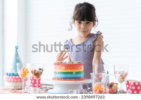 Caucasian little happy kid girl tasting cream from finger, smiling on birthday party with beautiful rainbow cake decorated with sugar candies. Colorful jelly in glasses on table. Looking at camera