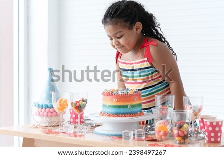 Smiling African little kid girl in cute dress, surprised on birthday party, looking at beautiful rainbow cake decorated with sugar candies. Colorful candies, jelly in glasses and hats on table