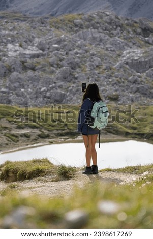 Young woman equipped with a backpack, taking pictures near a lake of the landscape with her mobile phone during a hiking route in the Dolomite Mountains