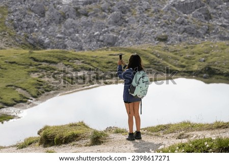 Young woman equipped with a backpack, taking pictures near a lake of the landscape with her mobile phone during a hiking route in the Dolomite Mountains