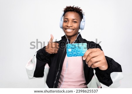 Delighted dark skinned black student guy wearing new modern headphones listening to music holding credit card and cell phone happy new bank customer.