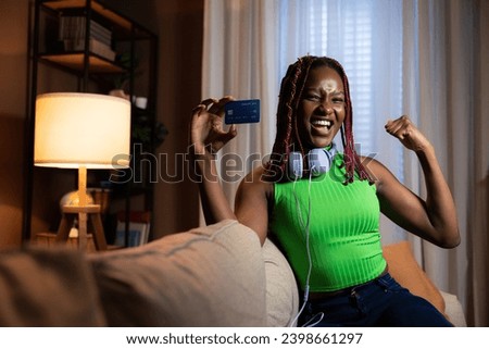 Photo of delighted cheerful afro american woman with colorful bright hair smiling looking at camera raising fist showing credit card at camera wearing modern new purple headphones sitting at home.