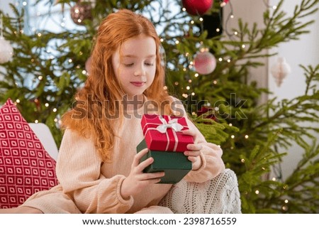 red-haired girls sister are looking at a box with gift against the backdrop of an interior with a Christmas tree.