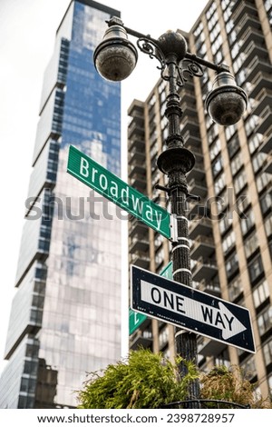 Broadway location direction and one way sign boards attached on signpost against blurred high rise buildings with glass walls in Manhattan, New York City during summer daytime under blue sky.