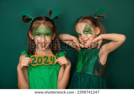 Funny sisters dressed up and made up in a green dragon, the symbol of the 2024 new year. Masked Children on a studio background