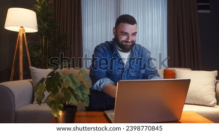 Young adult man sitting and relaxing on sofa at home using laptop enjoying surfing internet, using social media, chatting with friends