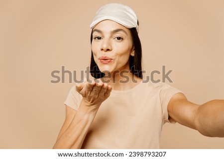 Close up young Latin woman wears pyjamas jam sleep eye mask rest relax at home do selfie shot on mobile cell phone blow air kiss isolated on plain beige background studio. Good mood night nap concept
