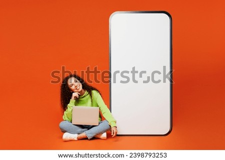 Full body young IT woman of African American ethnicity wear green hoody casual clothes big huge blank screen mobile cell phone with area use laptop pc computer isolated on plain red orange background
