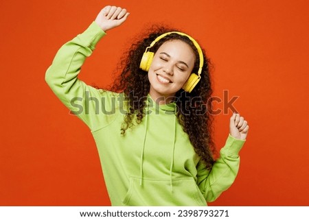 Young cheerful fun cool smiling woman of African American ethnicity she wear green hoody casual clothes listen to music in headphones dance isolated on plain red orange background. Lifestyle concept