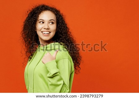 Side view young smiling fun woman of African American ethnicity she wear green hoody casual clothes point thumb finger aside on area mockup isolated on plain red orange background. Lifestyle concept