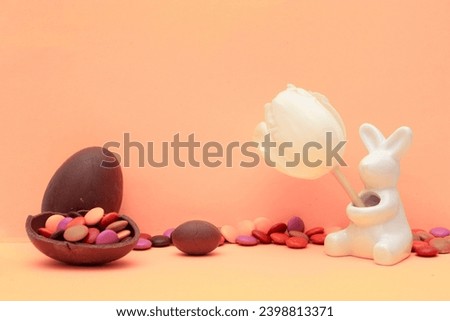 Sweets and ceramic bunny figurine on beige background. Food photo. Postcard for religious holiday Happy Easter. Chocolate eggs, candies and white rabbit with tulip. Trendy peach color of 2024